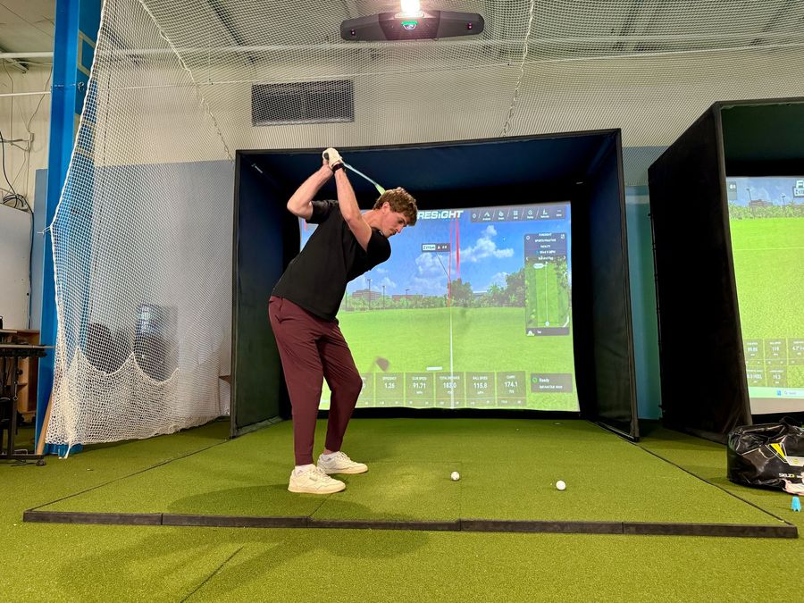 Man swinging golf club in simulator, wearing black shirt and burgundy pants.