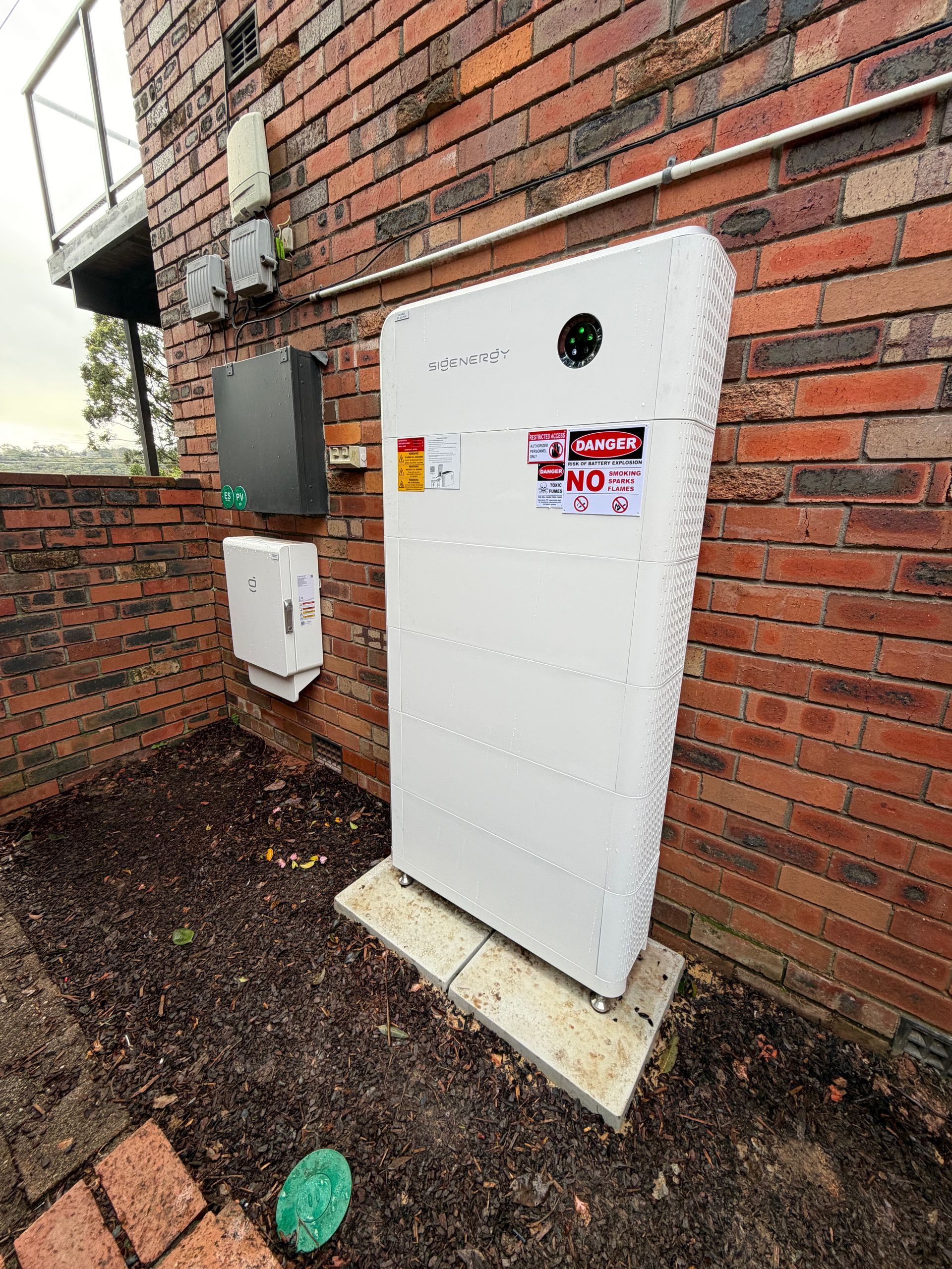White utility cabinet on a brick house exterior beside a smaller wall box and gravel ground