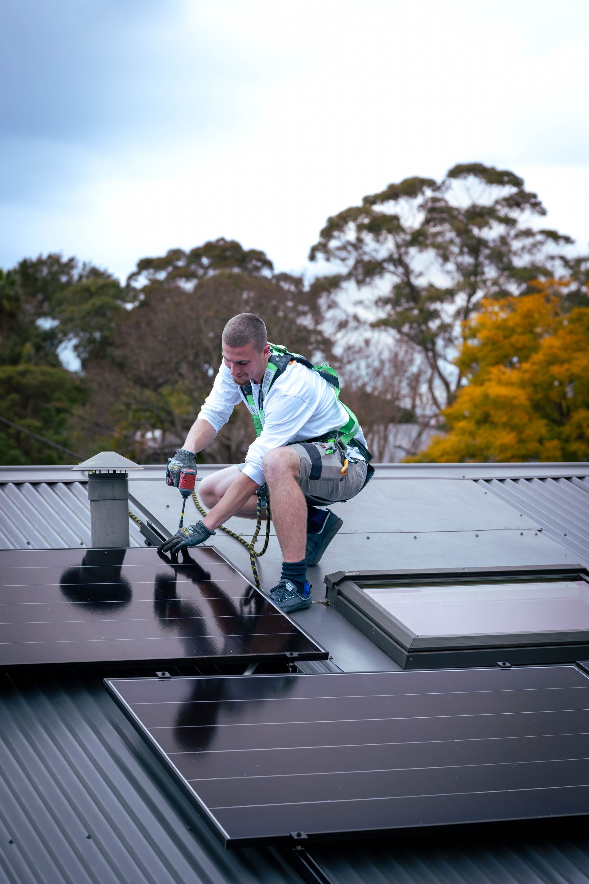 Worker crouching on a roof installing a solar panel, with trees in the background.