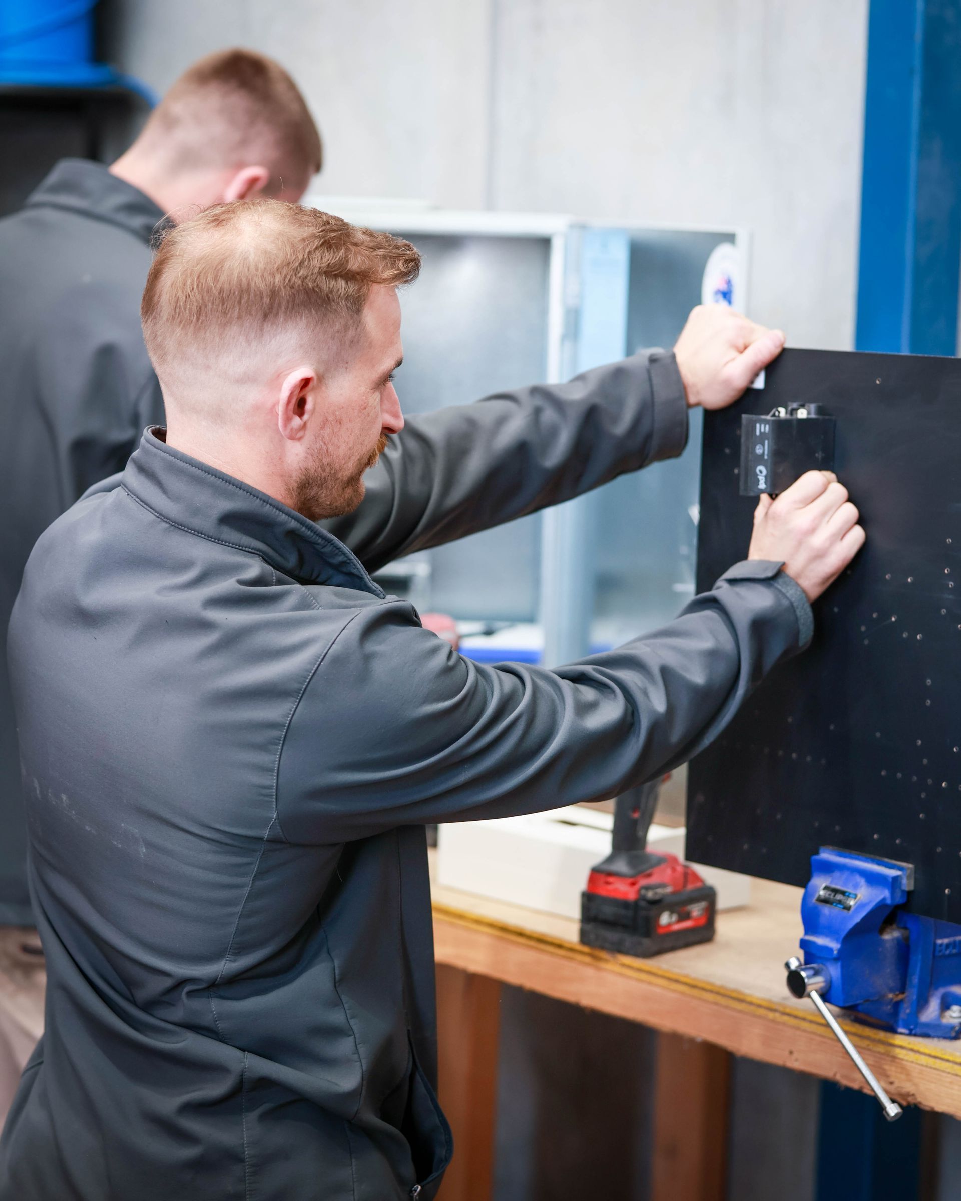 Two workers mounting a black panel in a workshop, one using a power drill at a workbench.