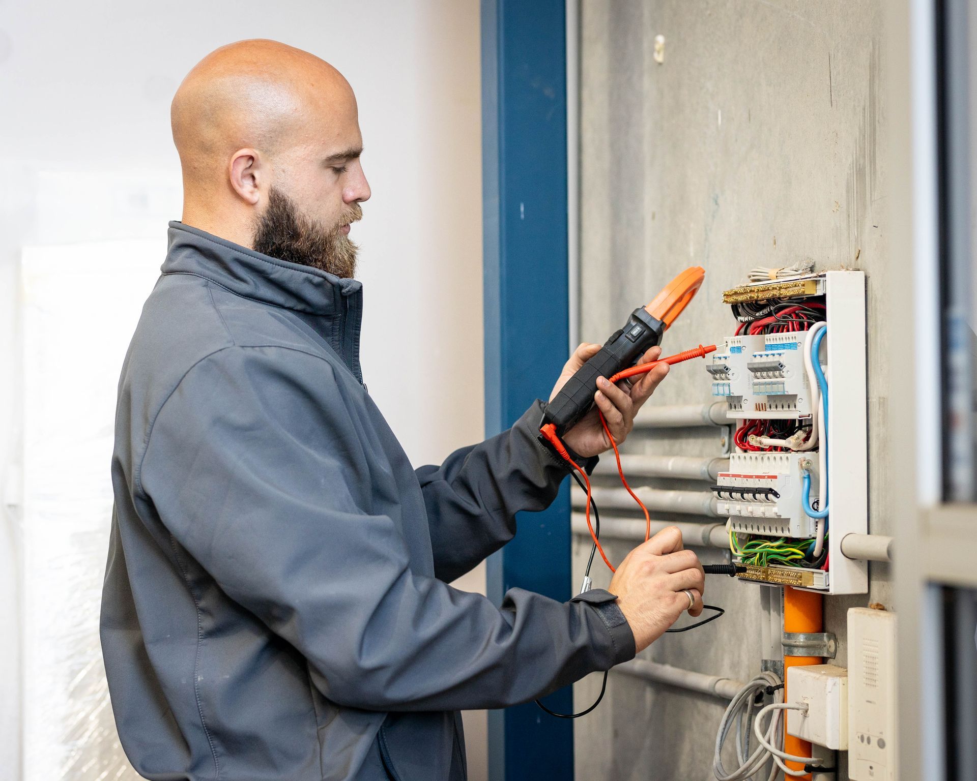 Electrician testing an open electrical panel with a multimeter, wearing a gray jacket and gloves.