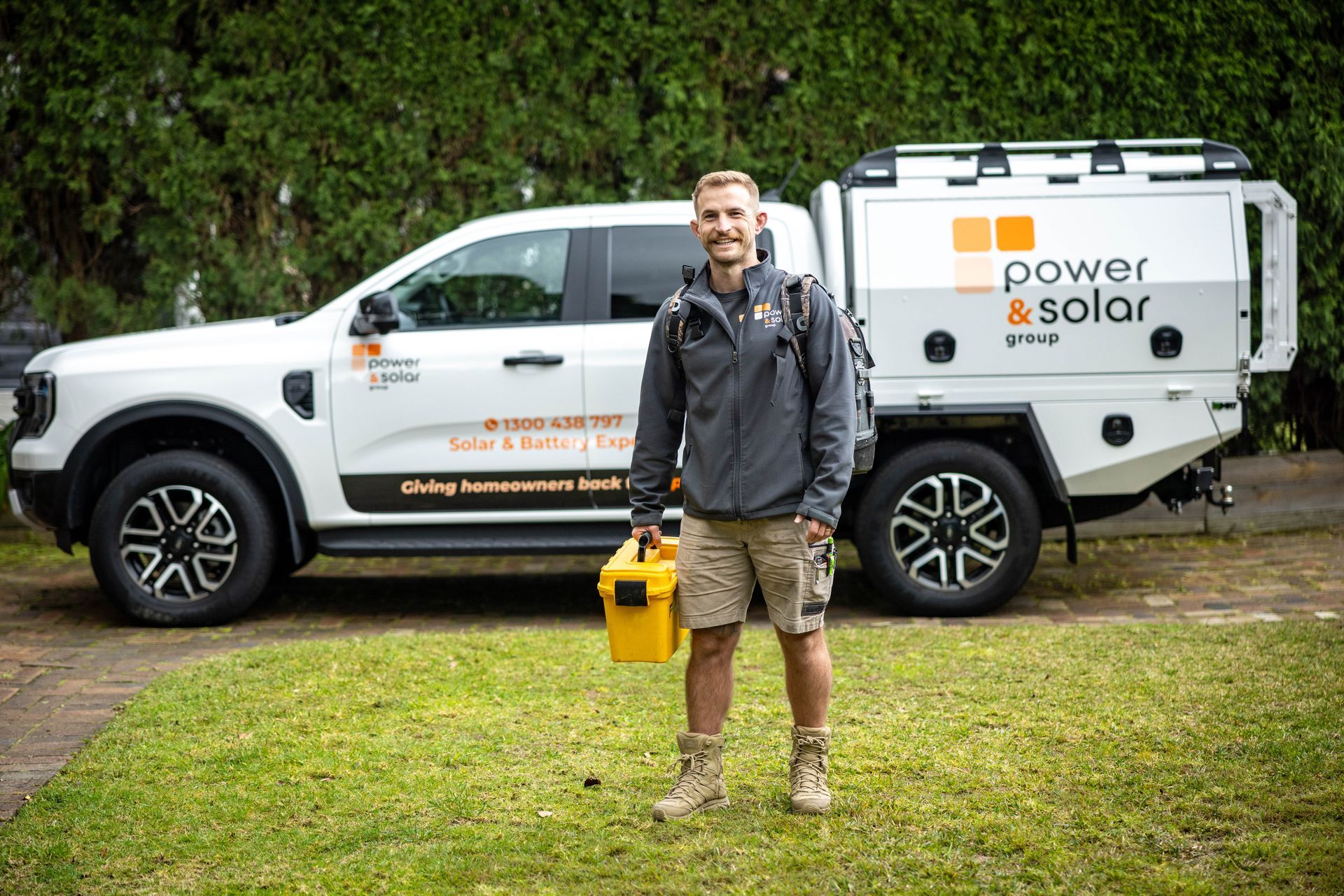Man standing beside a white Power Solar service truck on grass, holding a yellow toolbox.