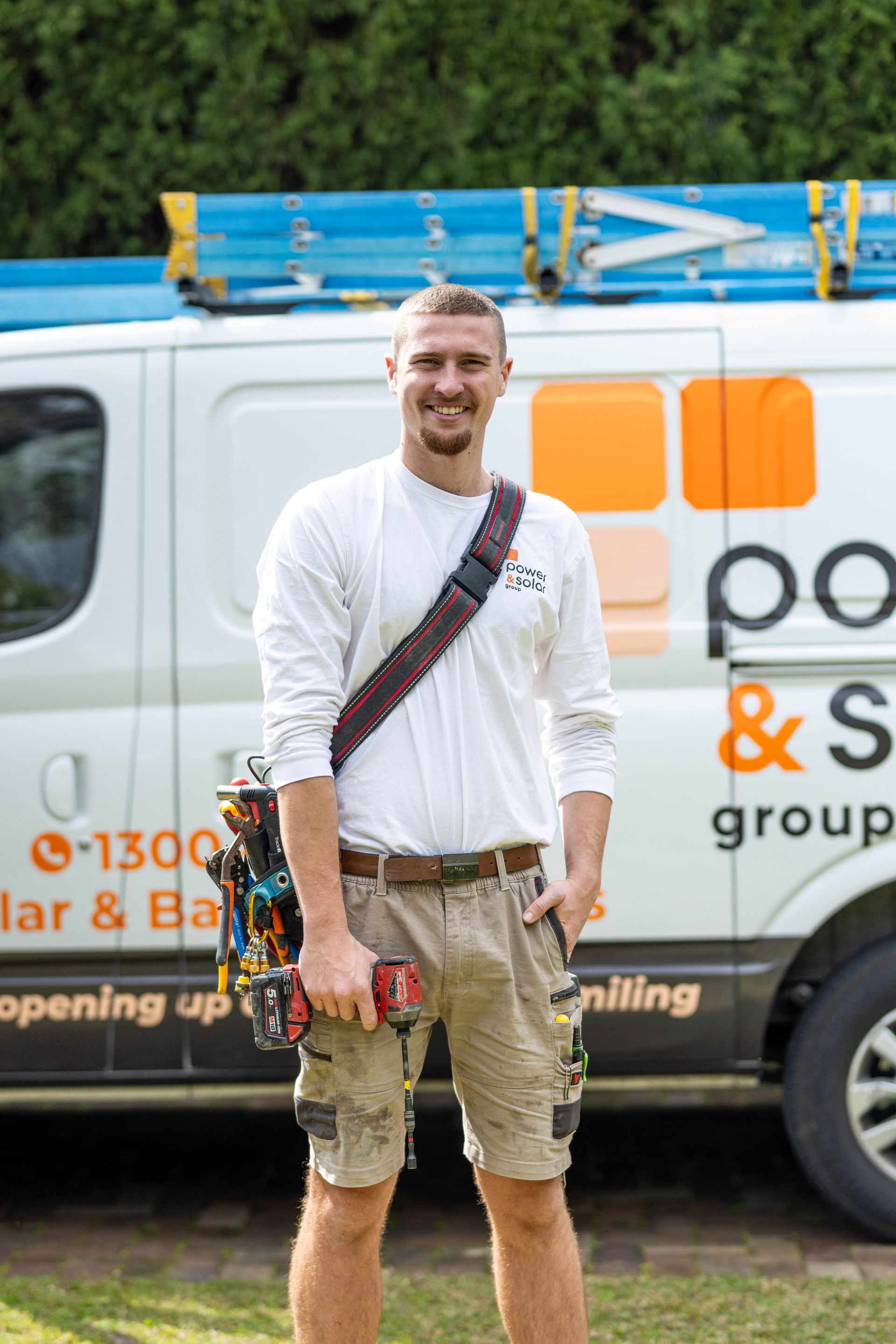 Smiling worker in white shirt and khaki shorts standing beside a service van with tools on a sunny day