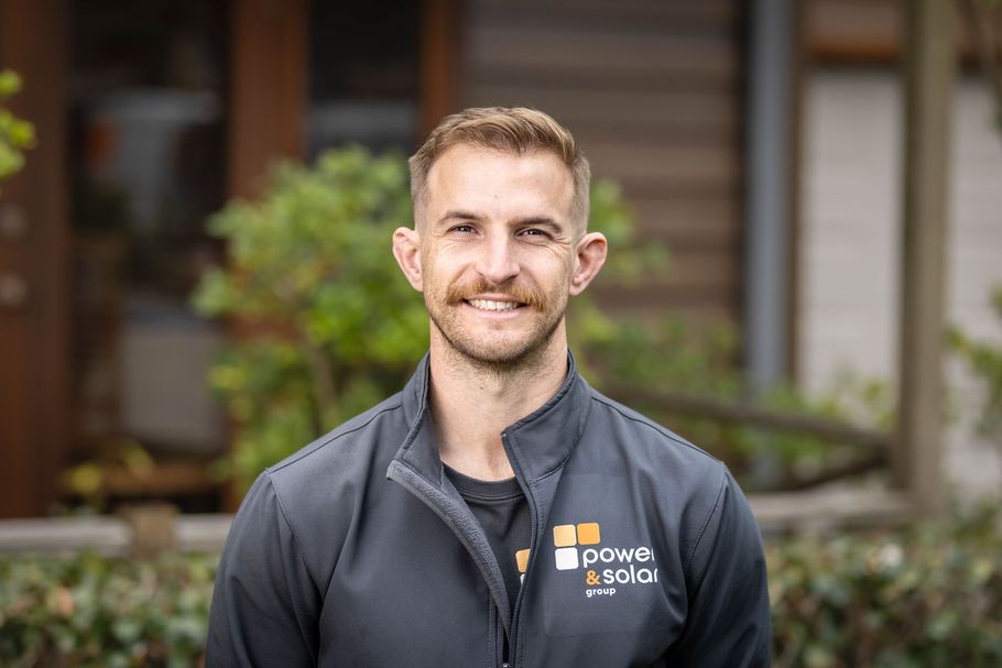 Smiling man in a dark jacket standing outdoors in front of a house and greenery