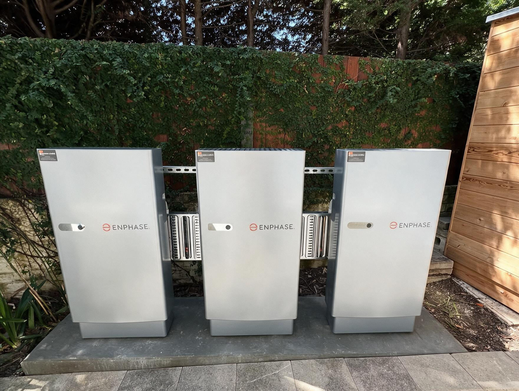 Three silver utility cabinets beside a hedge and wooden fence outdoors.