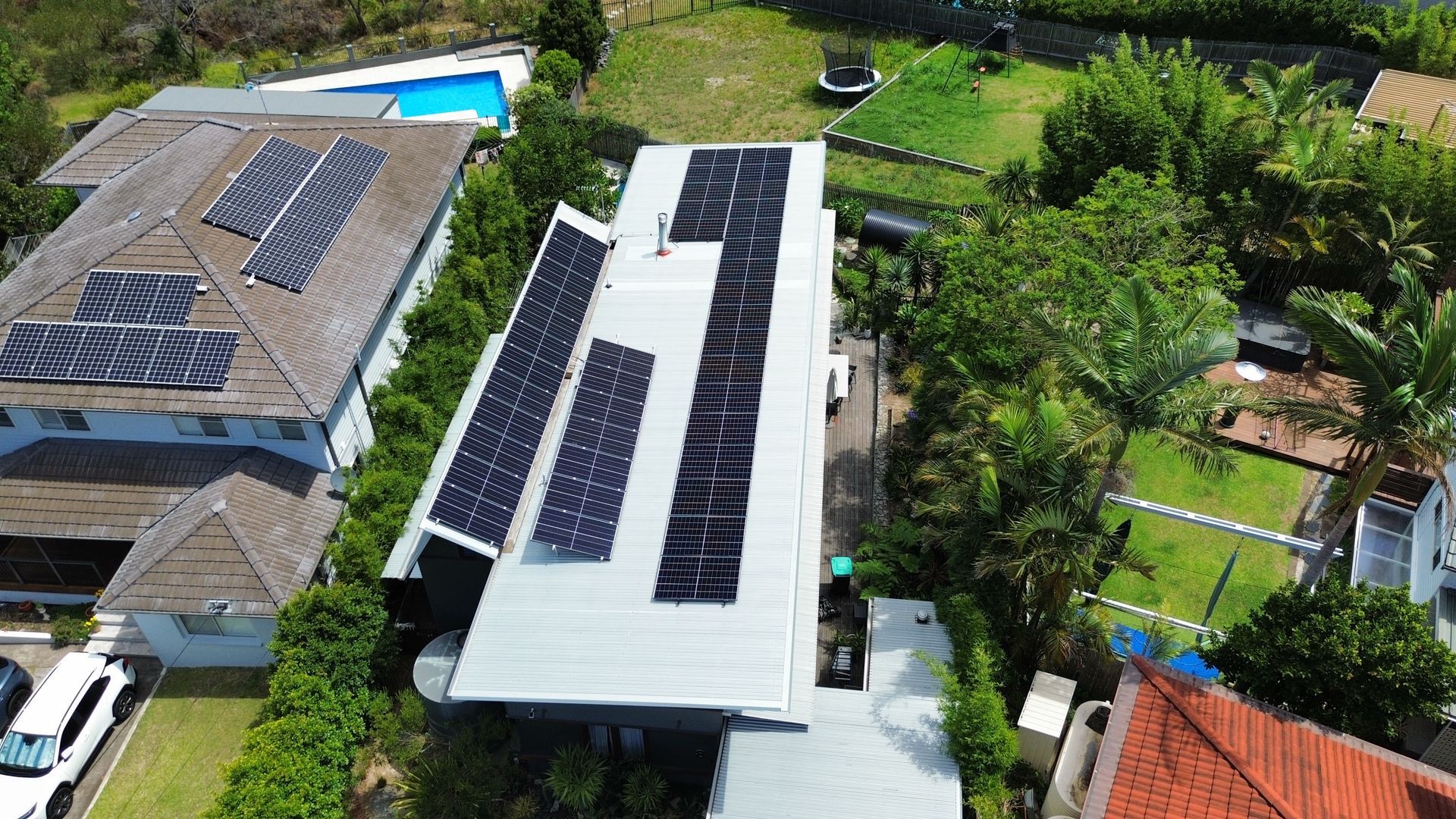 Aerial view of a house with rooftop solar panels beside a swimming pool and lush green trees.