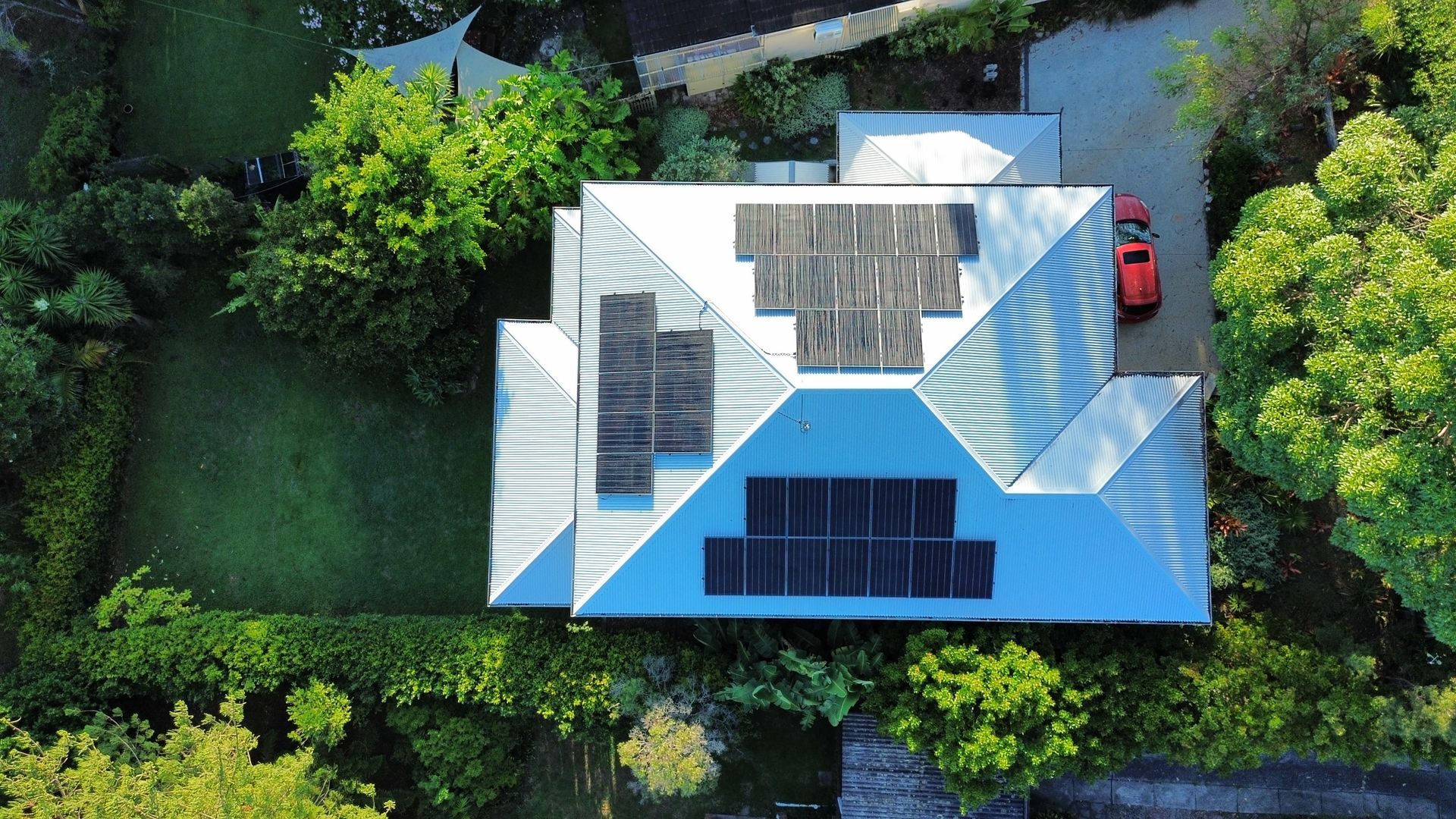 Aerial view of a house with a blue-and-white roof, surrounded by trees and a red car nearby.