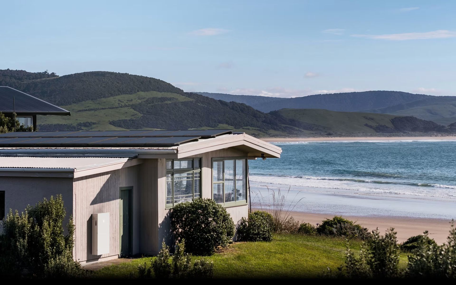 Coastal house overlooking a sandy beach and blue ocean, with green hills in the background