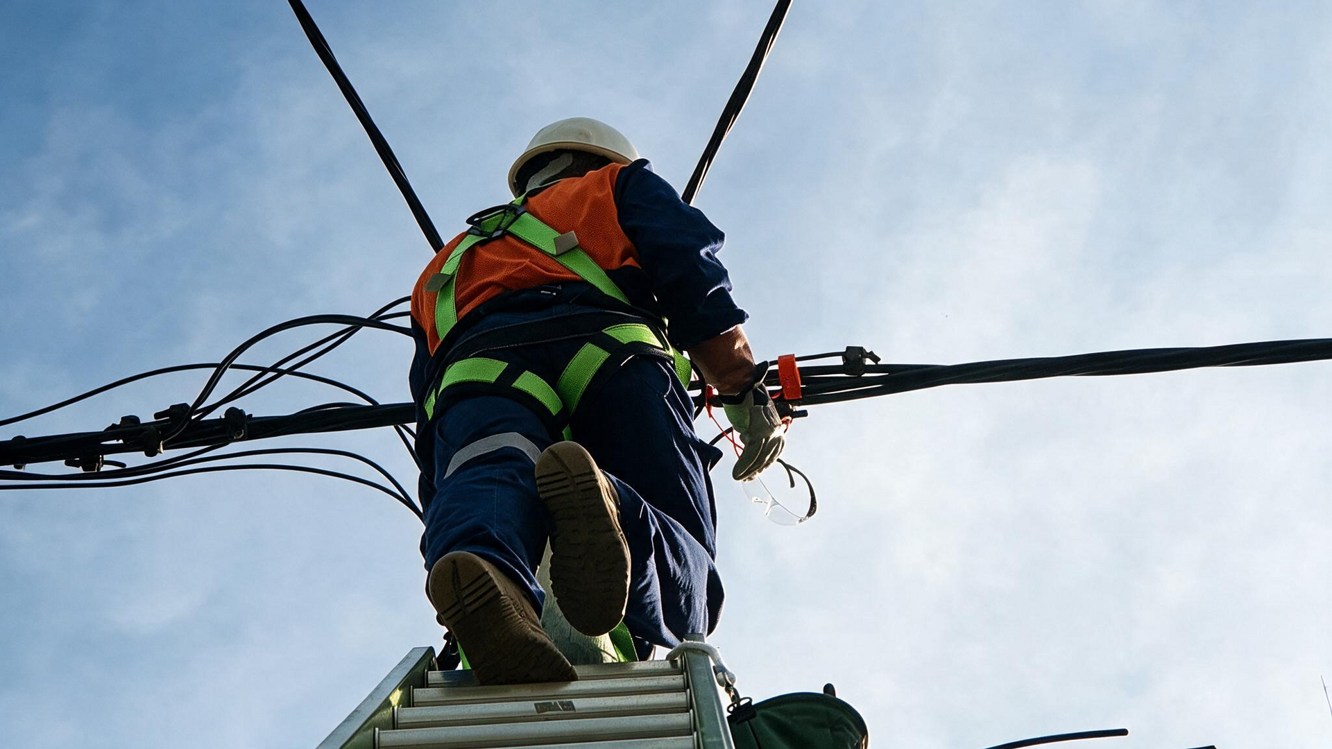 Utility worker in safety gear standing on a ladder, handling power lines against a blue sky