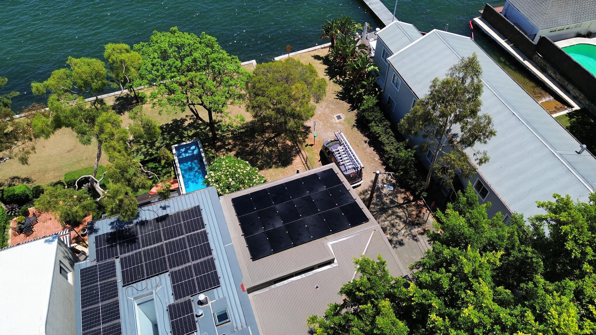 Aerial view of lakeside buildings with solar panels, trees, and a small pool beside the water