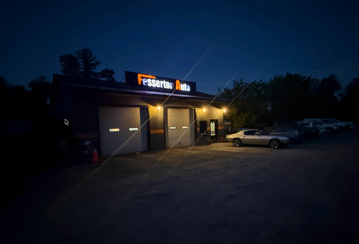 Nighttime view of a lit auto shop with parked cars in front and a glowing sign above the garage doors | Fesserton Auto