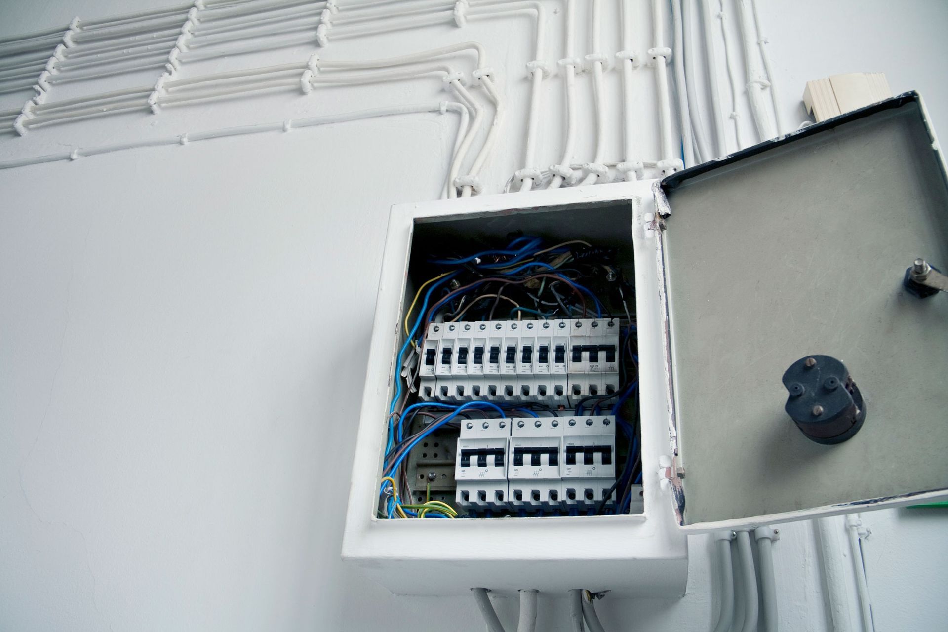 Open electrical panel with circuit breakers and wiring, mounted on a white wall, with conduit overhead.