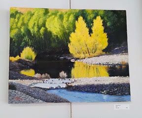 Painting of a river scene; yellow and green trees reflected in dark water with a stony shore.