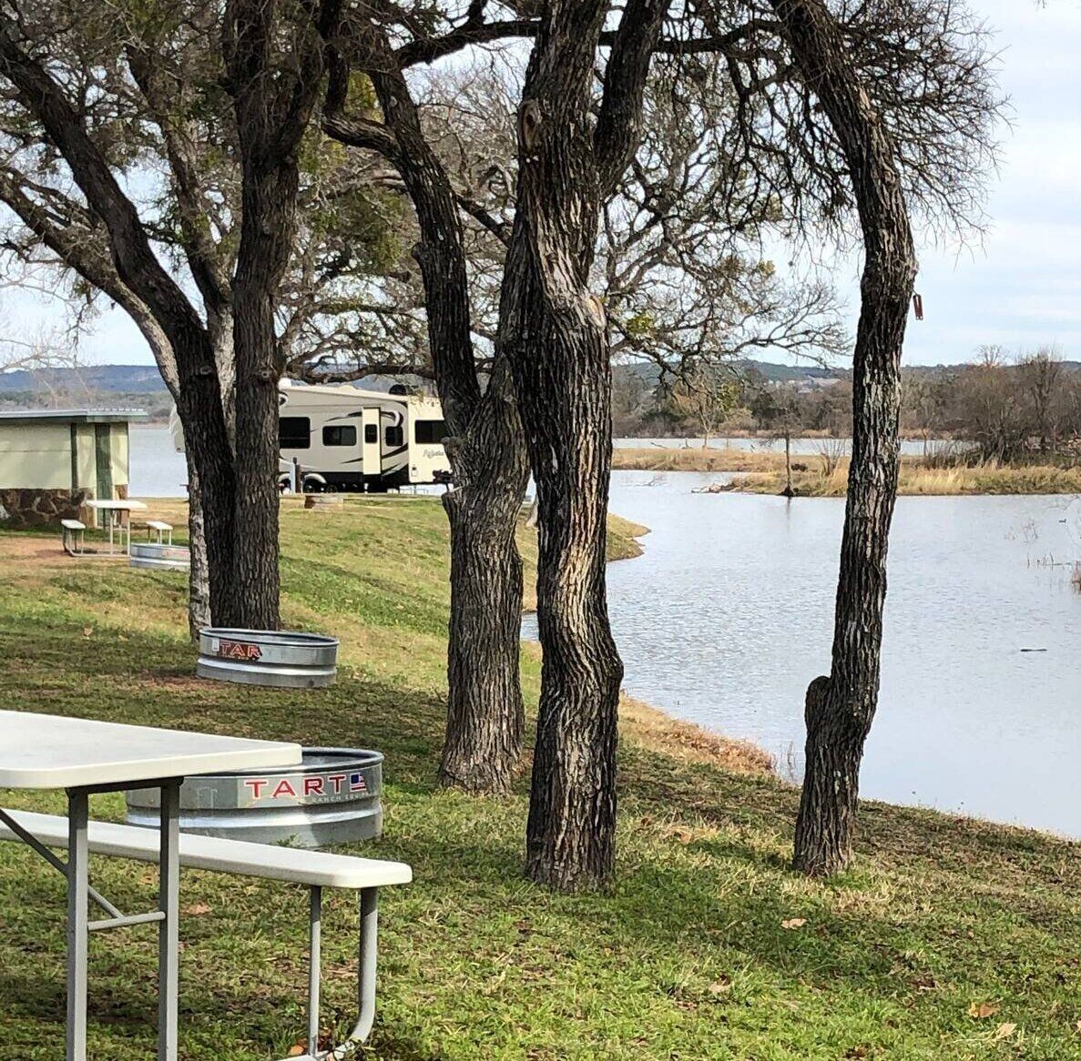 Riverside campsite with trees, picnic table, RV, and floodwater along the shore