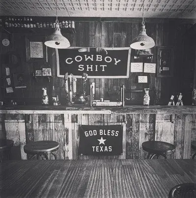 Rustic bar interior with a “Cowboy Shit” sign and a “God Bless Texas” banner behind the counter