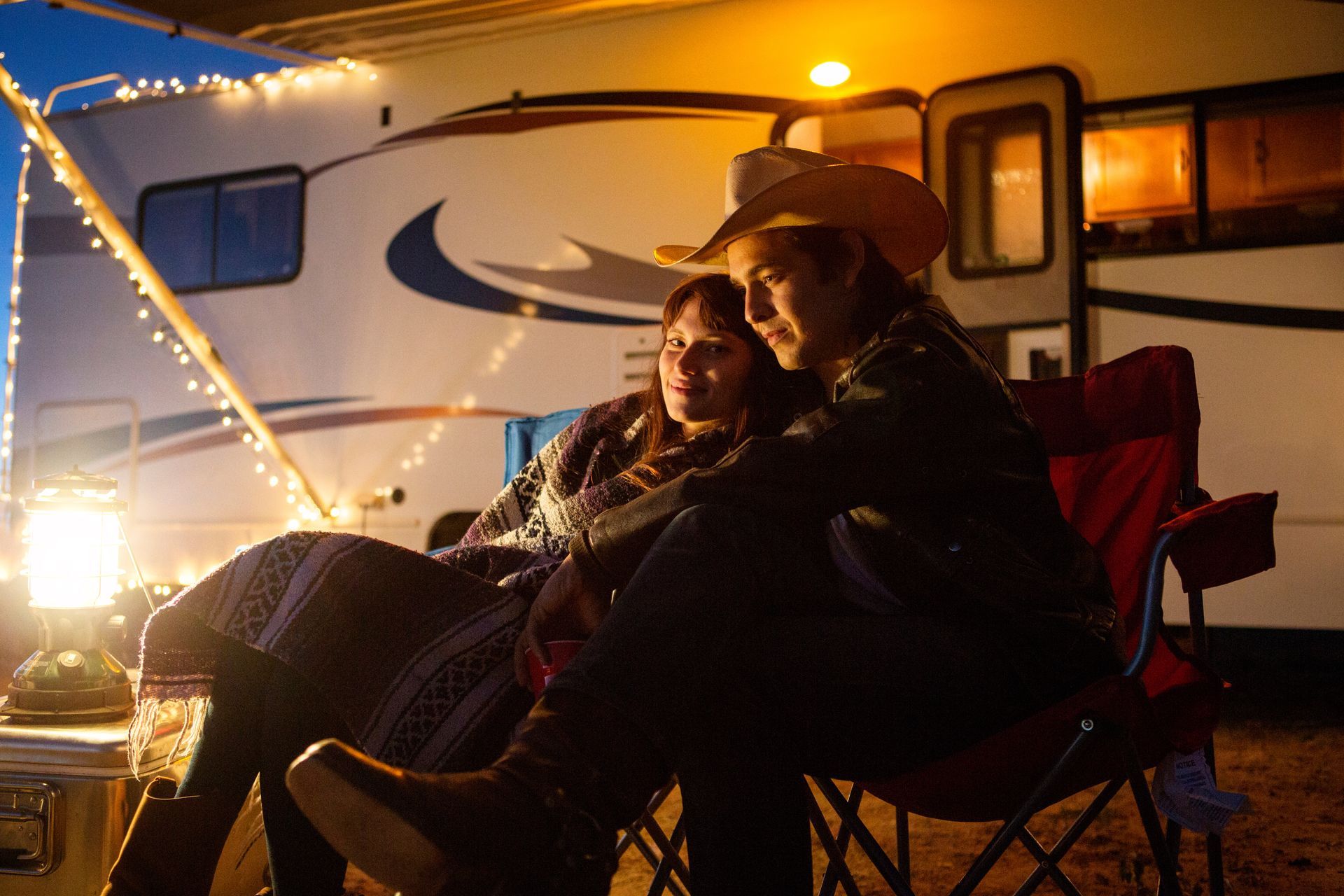 Couple sitting in folding chairs by an RV at night, lit by warm string lights.