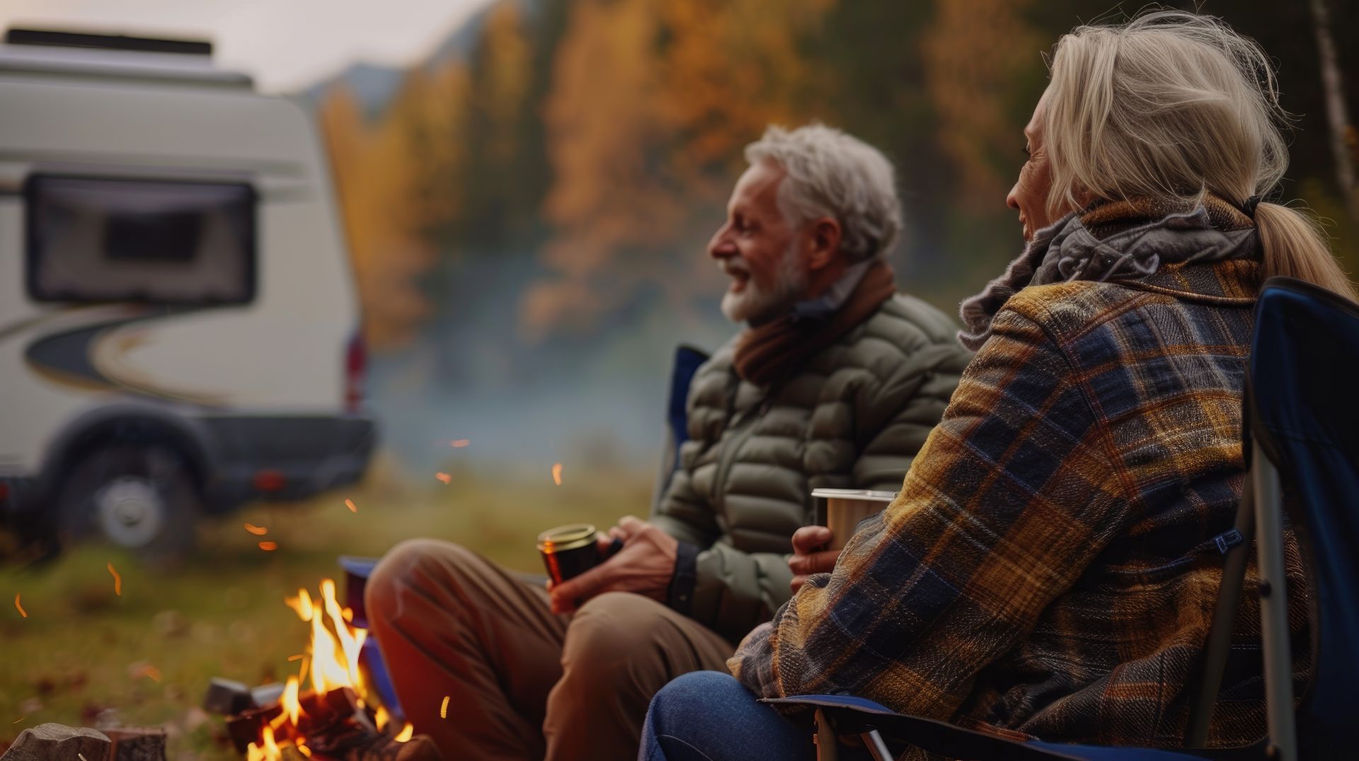 Two campers sit by a campfire beside an RV in a forest at dusk, wrapped in warm jackets.