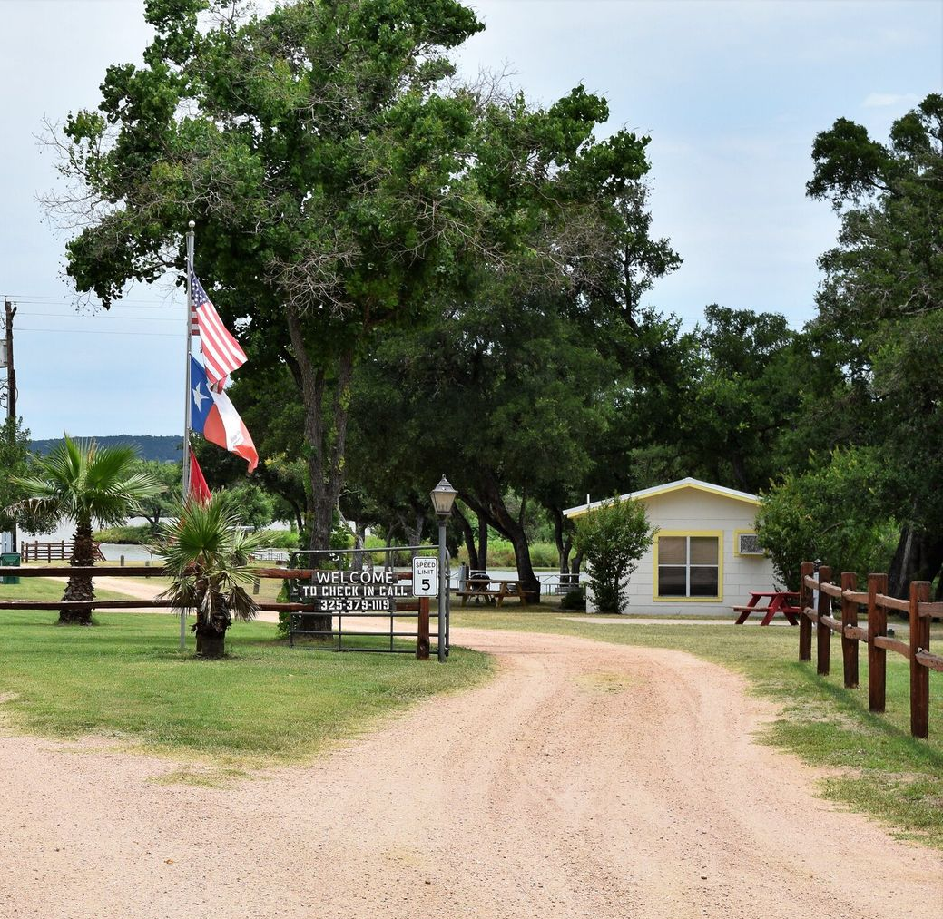 Dirt path leading to a small building beside trees and a pond, with Thai and U.S. flags on the left.