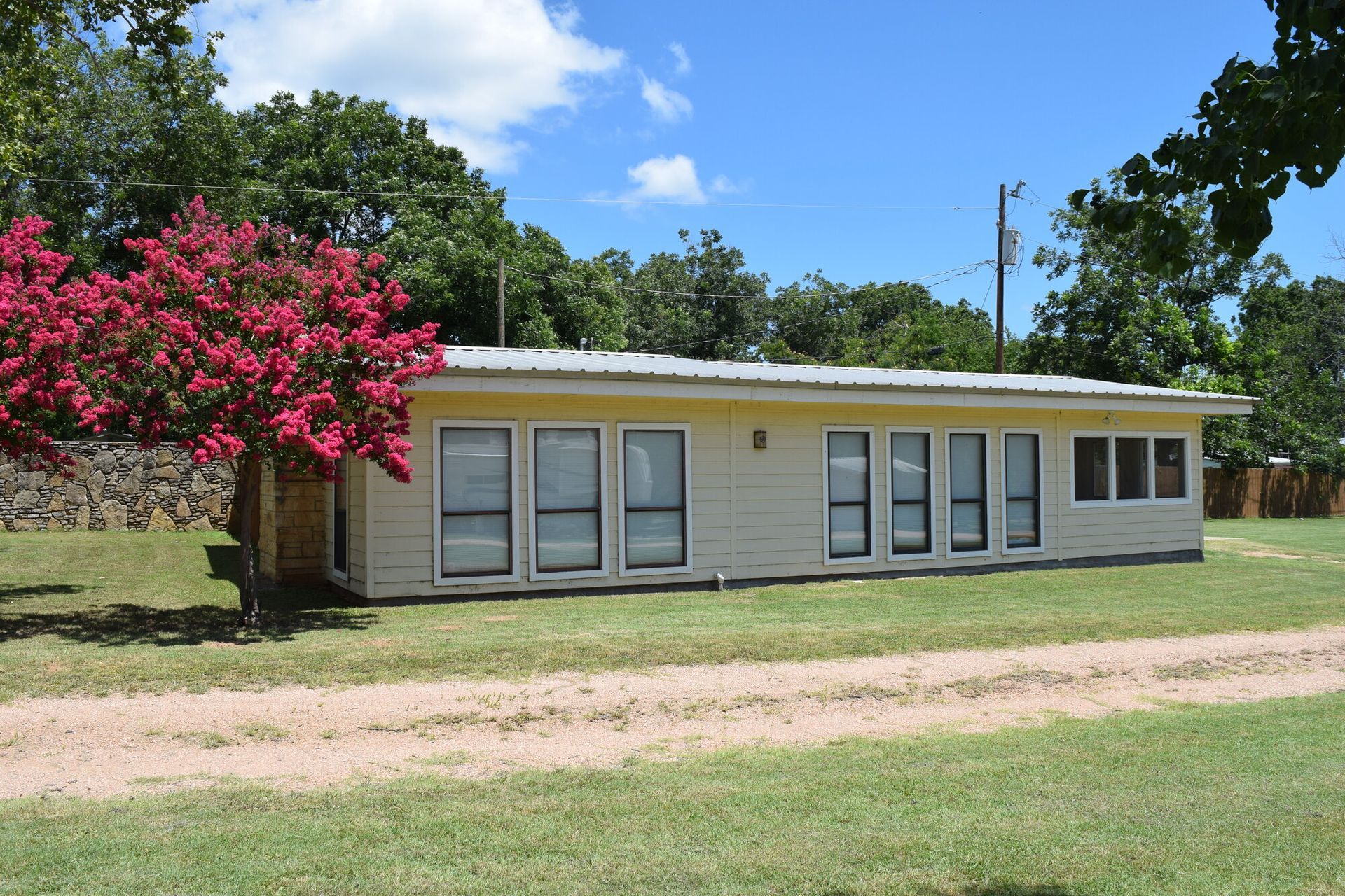 Single-story beige house with a white roof, pink flowering tree, and grassy yard under a blue sky