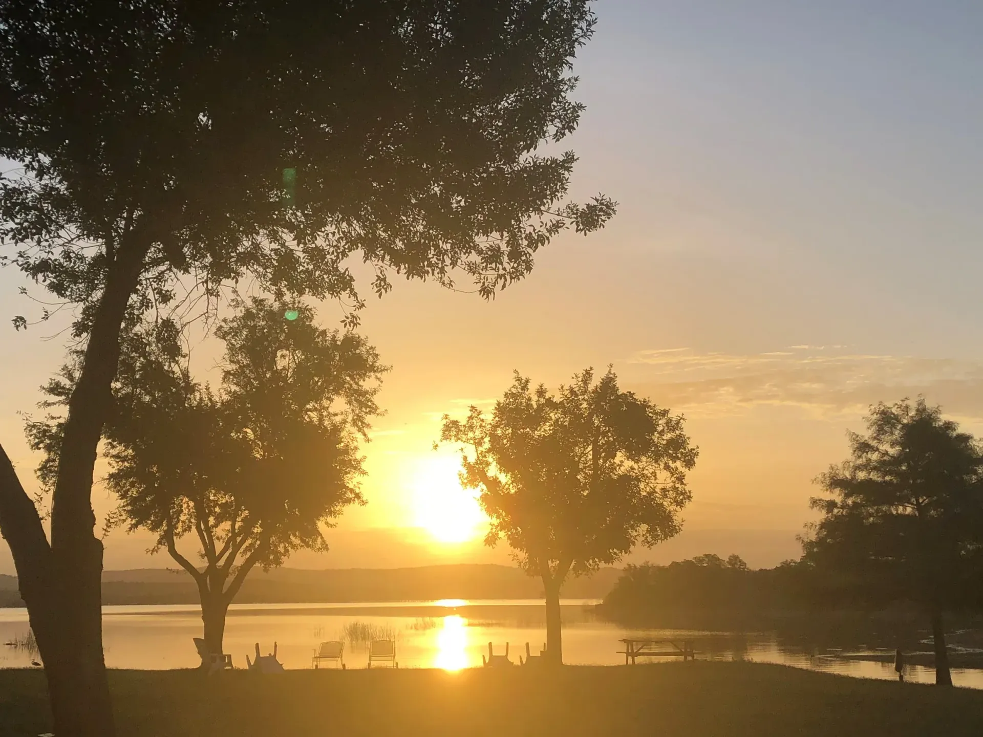 Sunset over a calm lakeside with silhouetted trees and a glowing reflection on the water