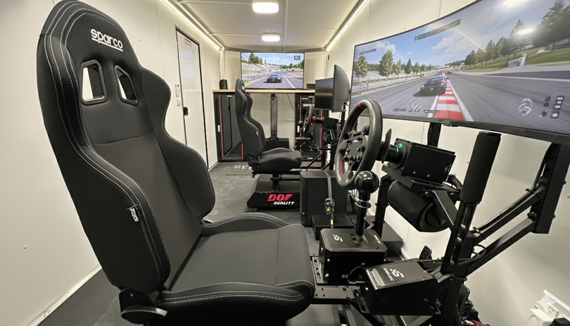 Racing simulator cockpit with a black seat, steering wheel, pedals, and a large screen showing a track view