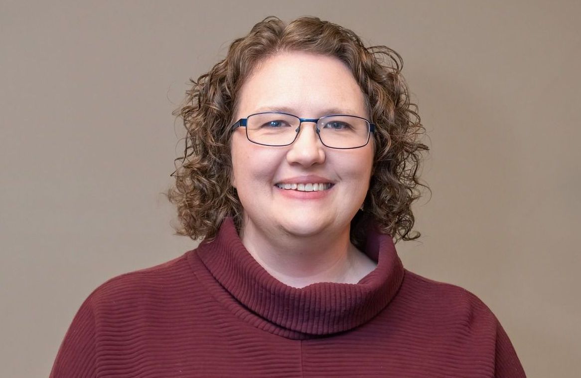 Smiling person with curly hair and glasses in a maroon sweater against a neutral background