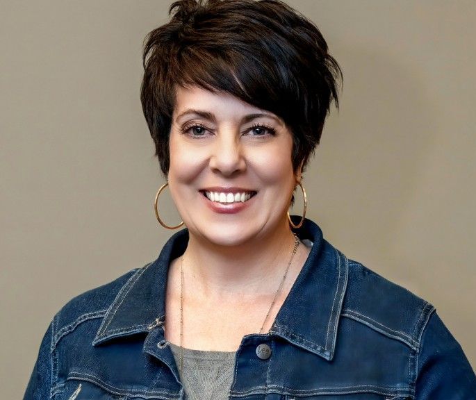 Smiling woman with short dark hair wearing a denim jacket and hoop earrings against a plain background