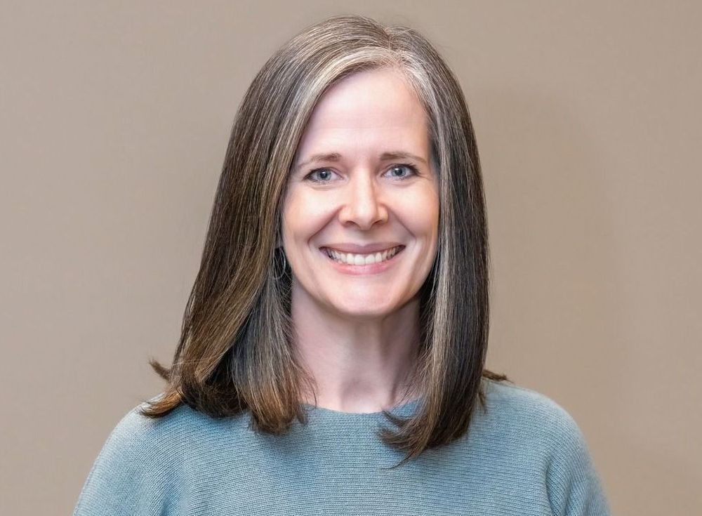 Smiling woman with shoulder-length hair wearing a gray sweater against a beige background