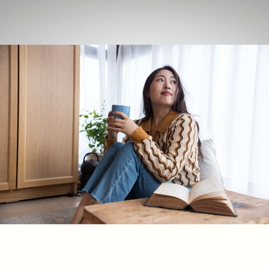 Woman sitting by a window, holding a mug beside an open book in a cozy room