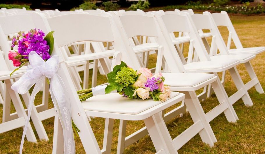 White folding chairs decorated with purple and pink flowers arranged outdoors on grass for a ceremony