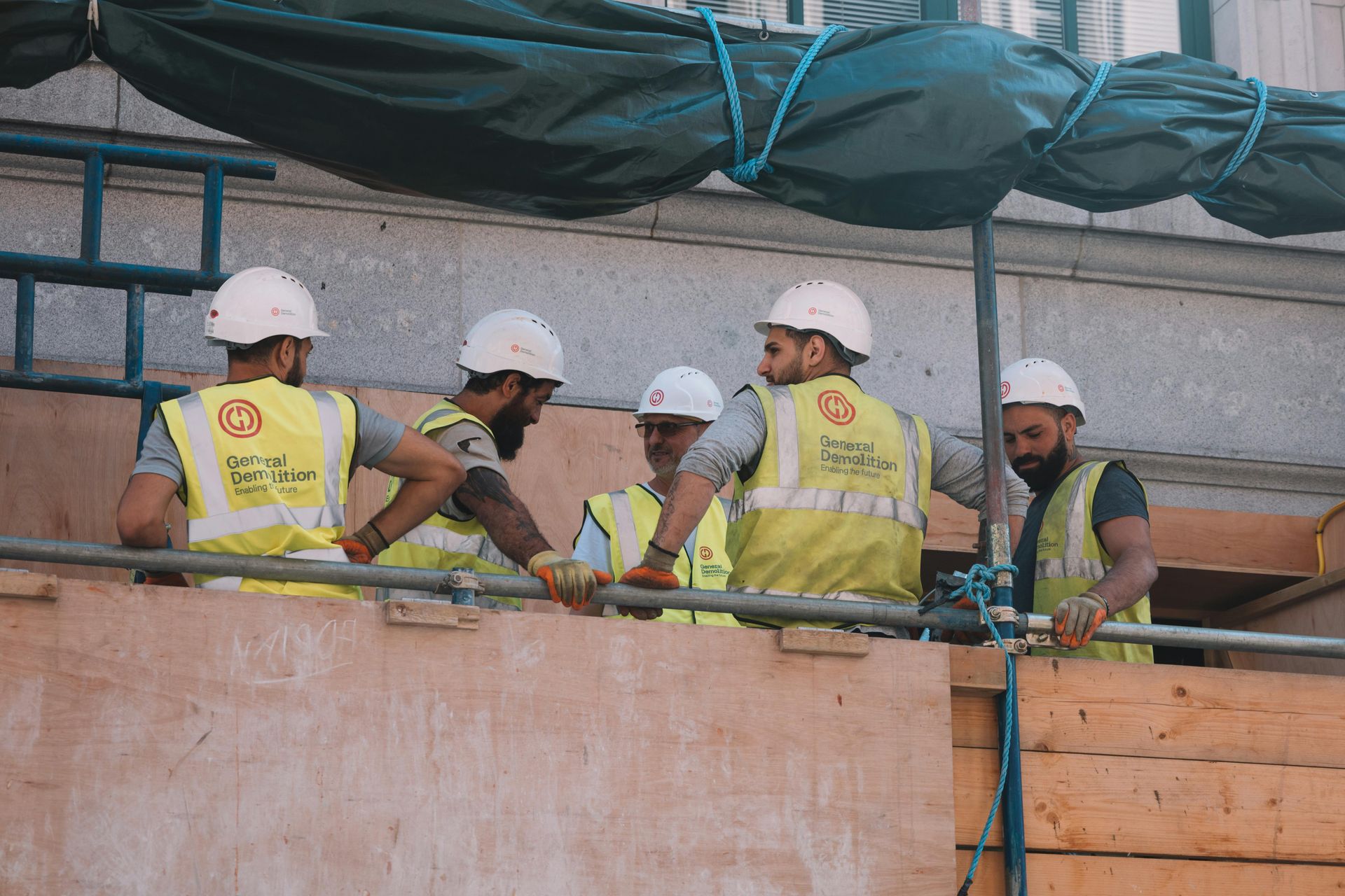Five construction workers in hard hats and reflective vests work on a scaffold behind a barrier.