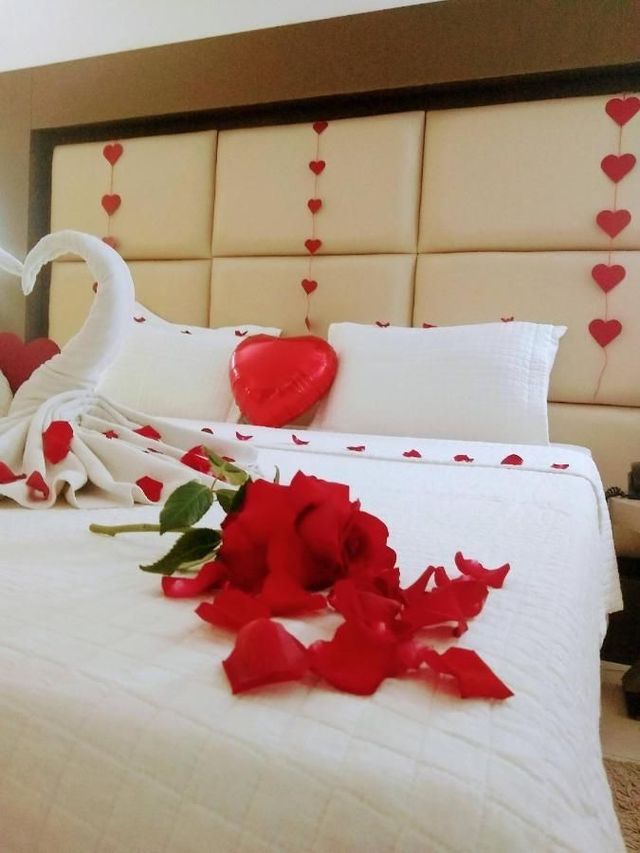 Quarto romântico decorado com pétalas de rosa, corações e uma toalha em formato de cisne sobre uma cama branca.