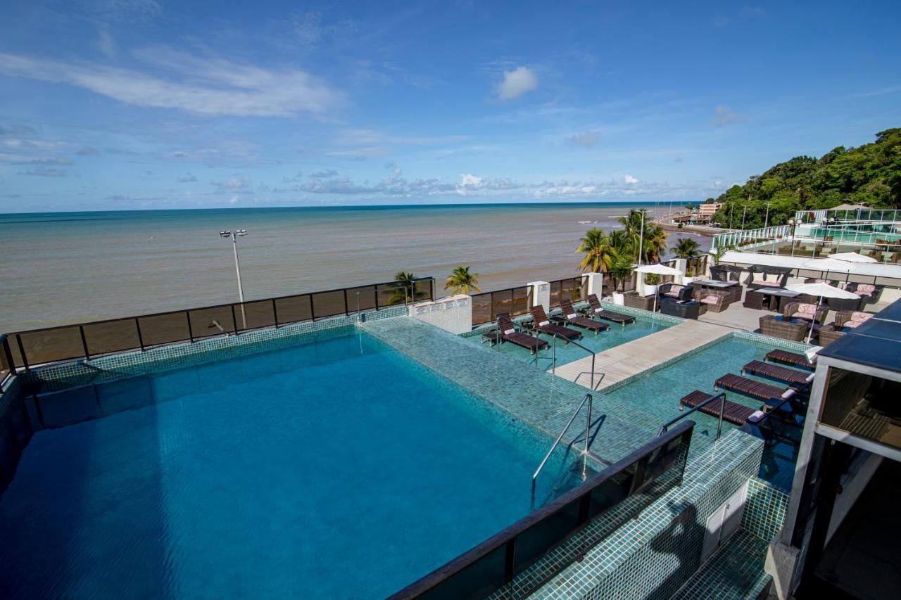 Piscina com vista para o oceano sob um céu azul; espreguiçadeiras, palmeiras.