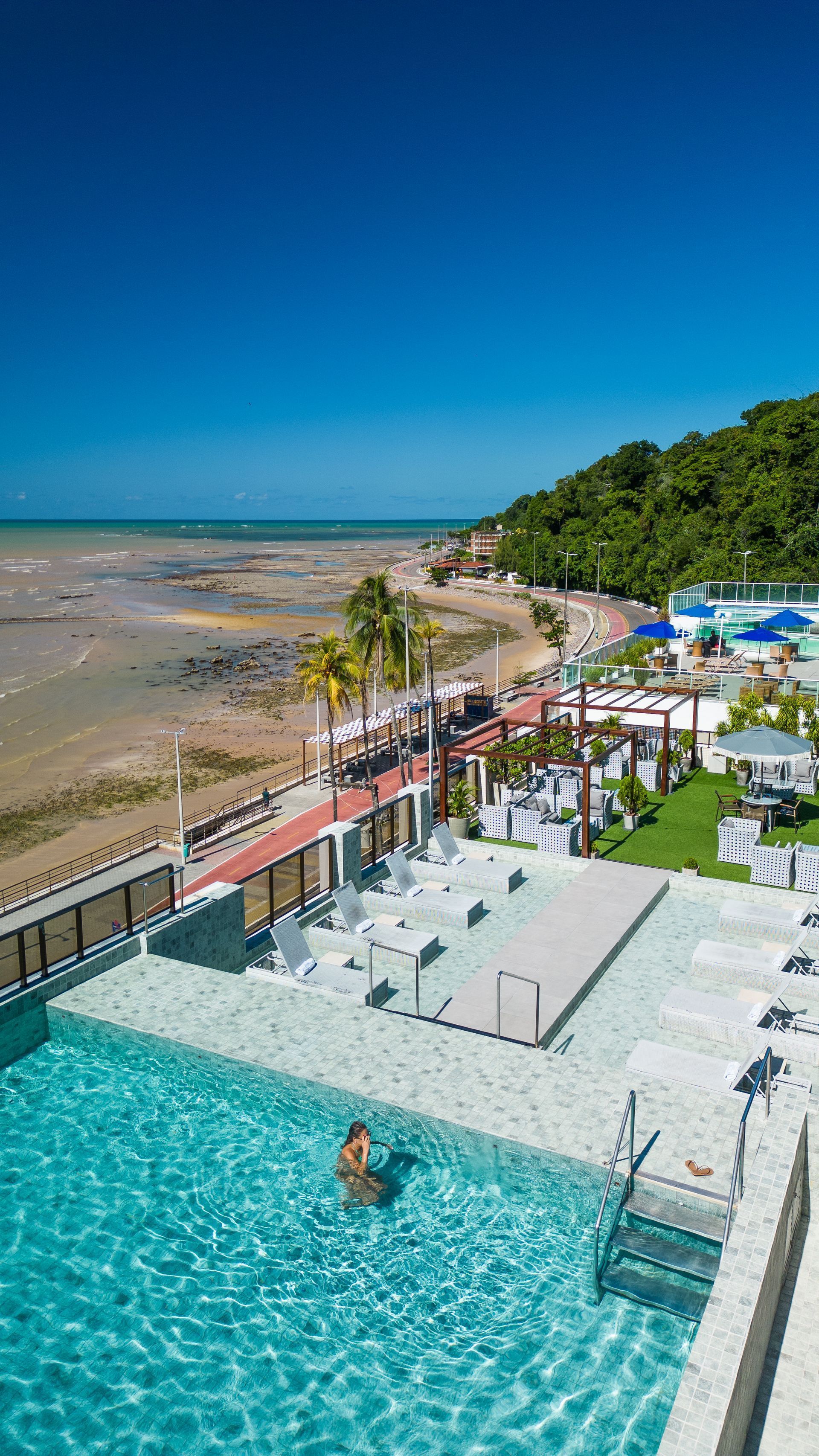 Piscina infinita com vista para a praia e o oceano. Uma pessoa nada na piscina. Dia ensolarado com céu azul.