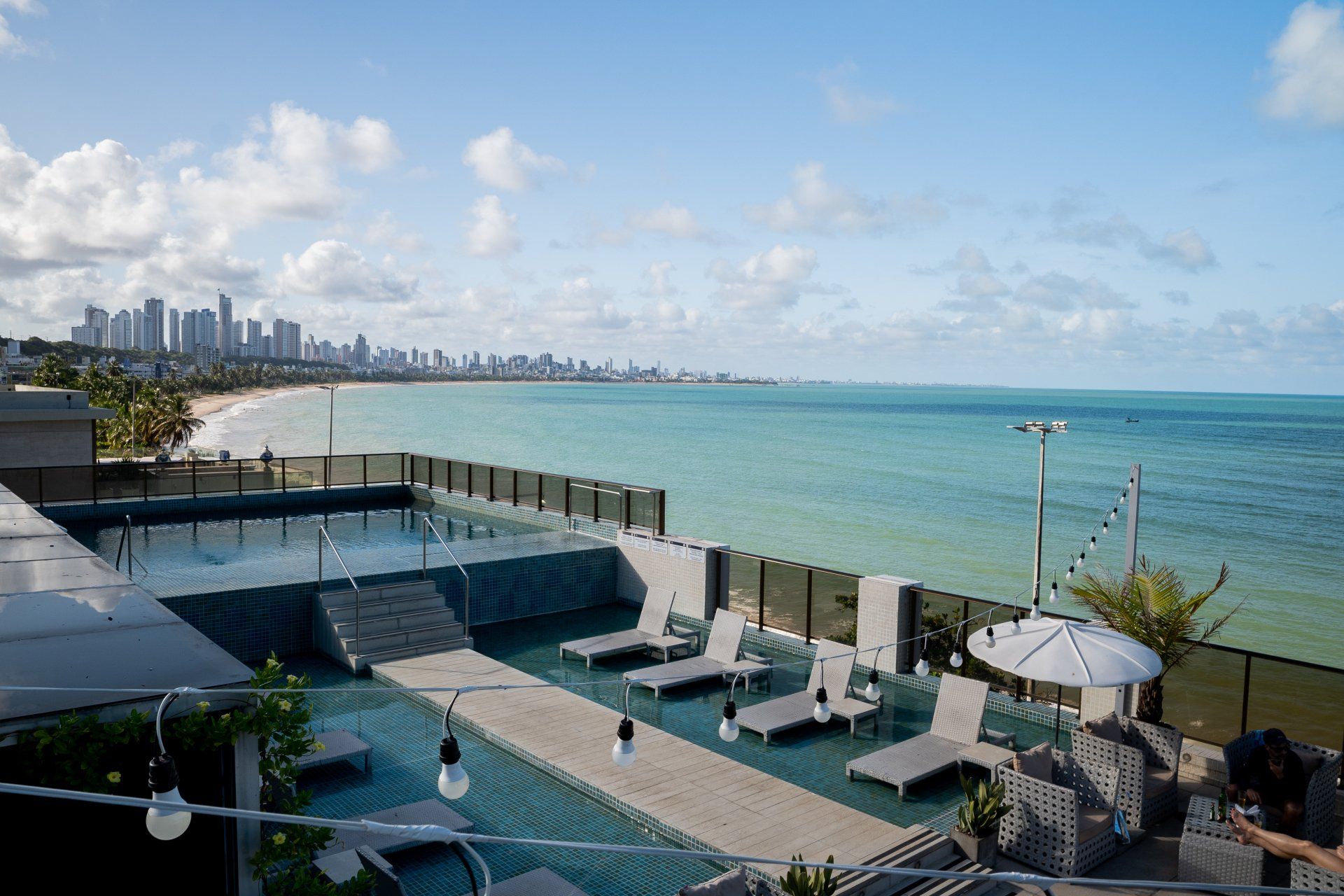 Piscina na cobertura com vista para a praia e para o horizonte da cidade em um dia ensolarado. Cadeiras de descanso e luzes de corda visíveis.