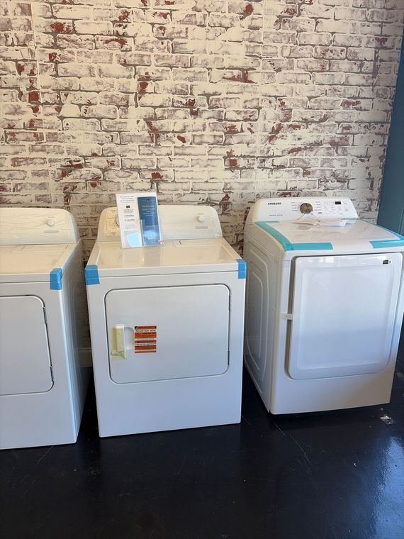 Three laundry appliances lined up against a brick wall on a dark floor.