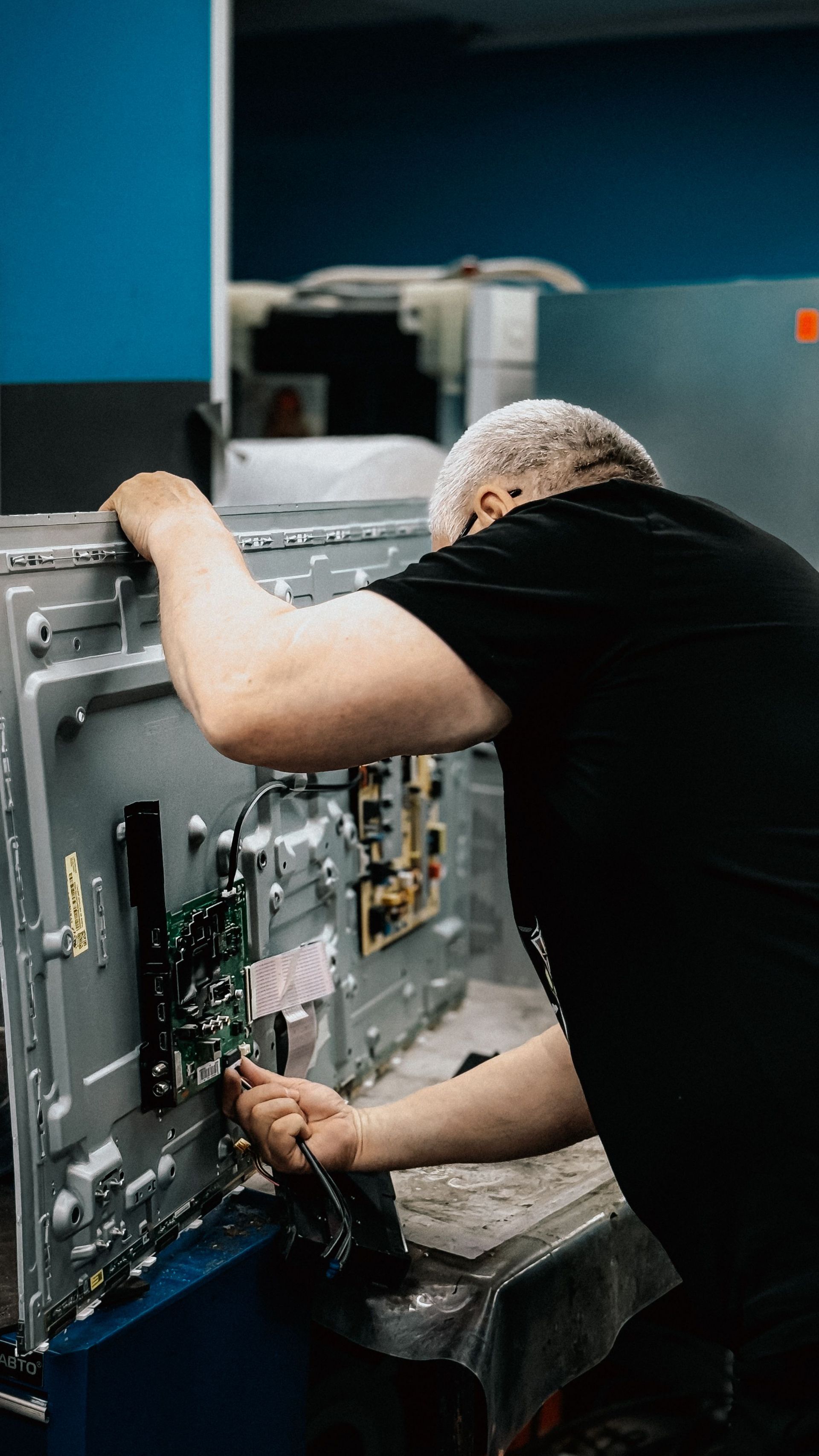 Technician repairing the back of a large flat-screen TV in a workshop, with tools and exposed circuitry.