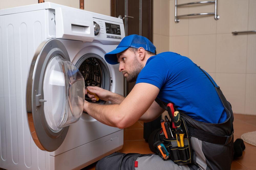 Technician in blue cap repairs a front-loading washing machine with tools in a laundry room