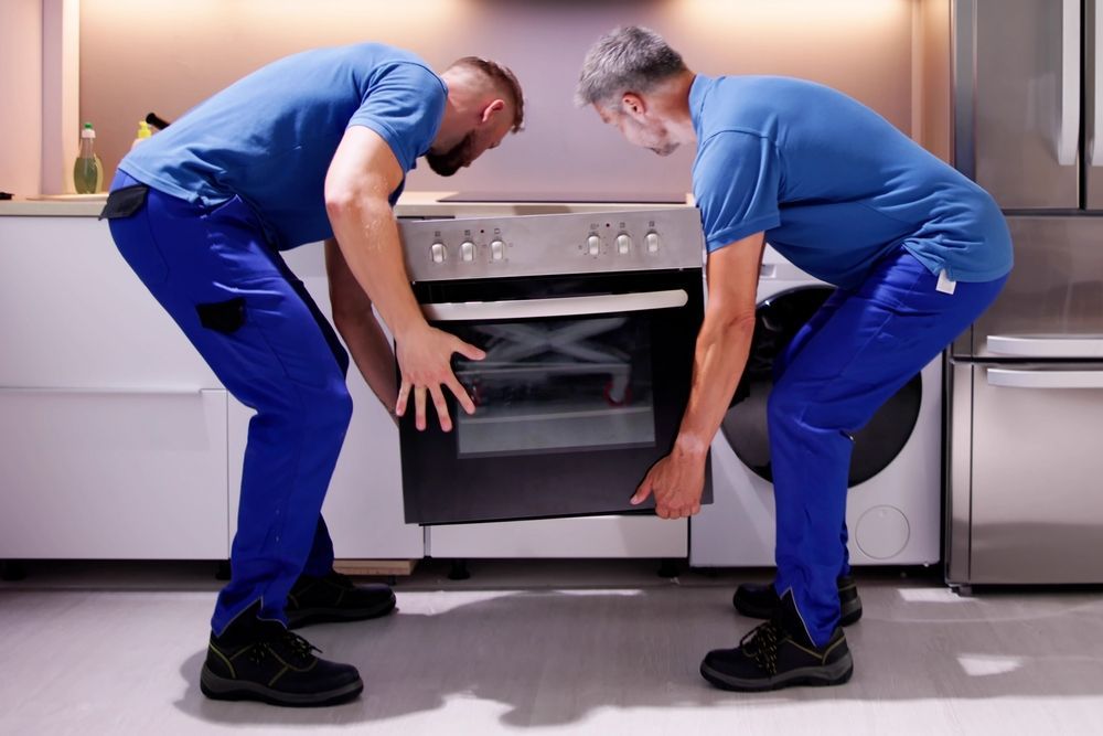 Two workers in blue uniforms lifting a stainless steel oven into place in a modern kitchen