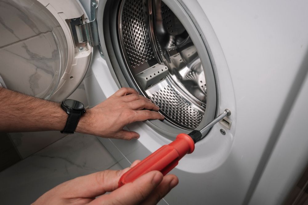 Hands using a red screwdriver to repair a front-loading washing machine door hinge