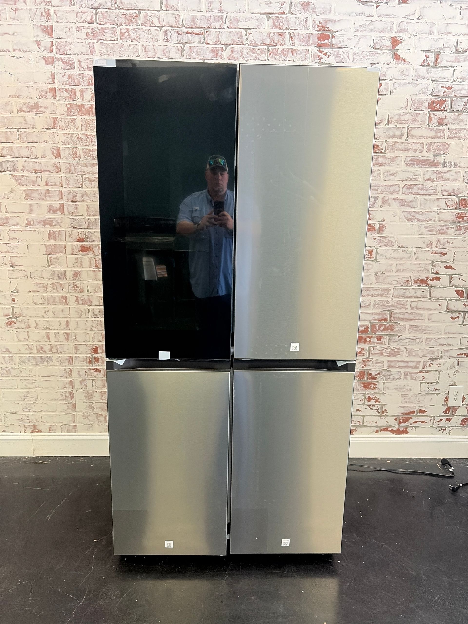 Four-panel refrigerator with black and silver doors against a white brick wall