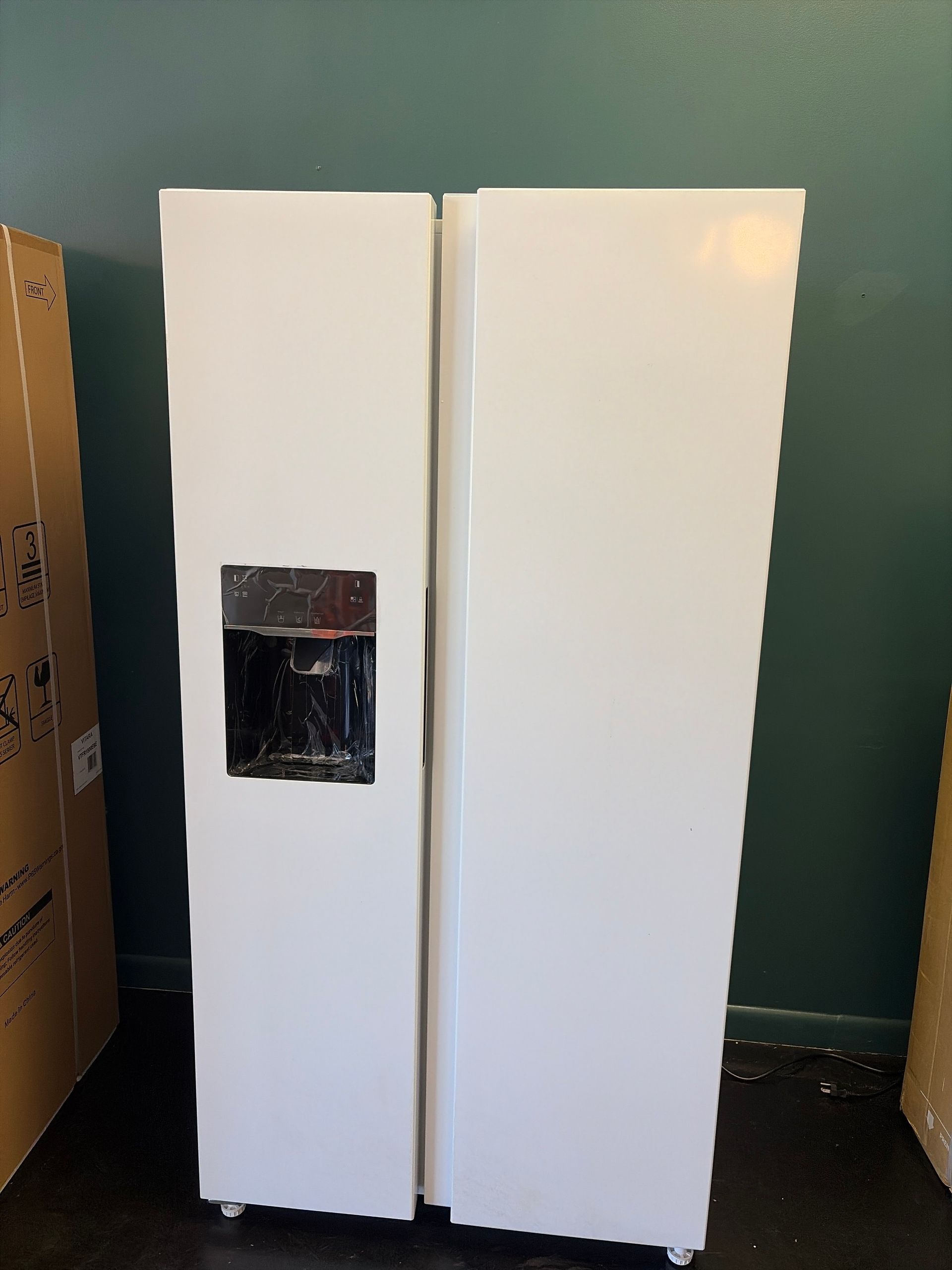 Two white refrigerators side by side, one with a small black dispenser panel, against a green wall.