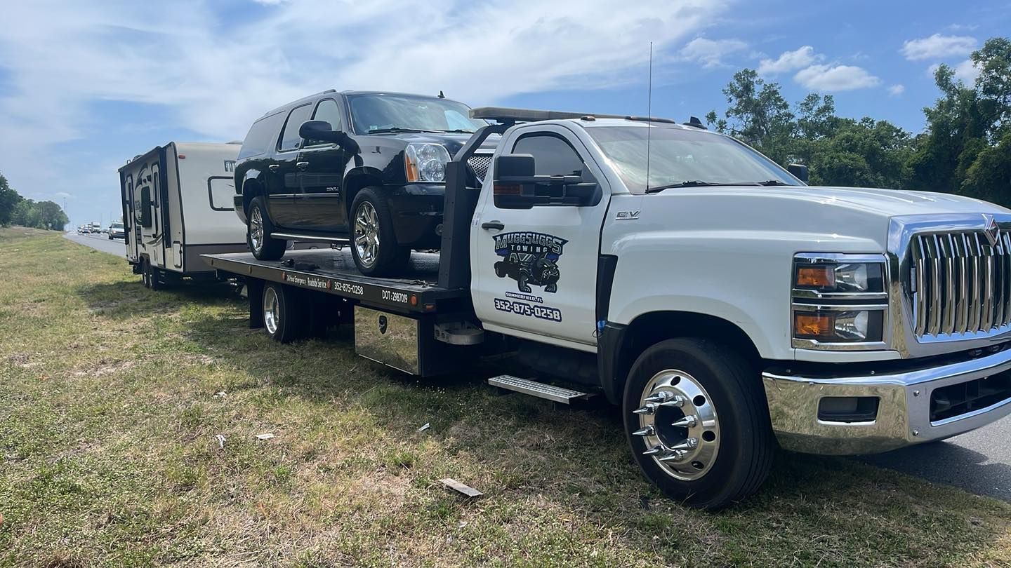 Tow truck hauling a damaged black SUV on a flatbed in a grassy field under a blue sky