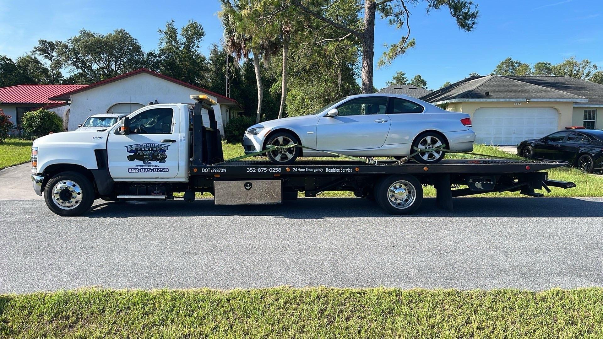 A white tow truck with a silver car on its flatbed, parked on a suburban street.