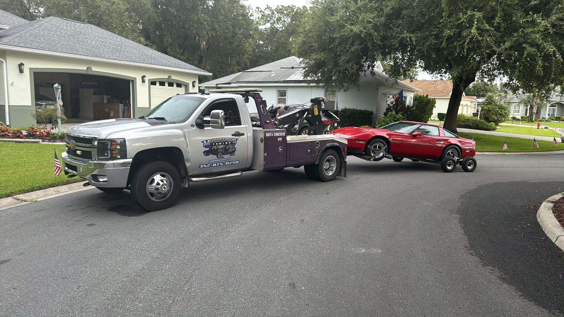 Tow truck towing a red car on a residential street. The tow truck is silver, the sky is overcast.