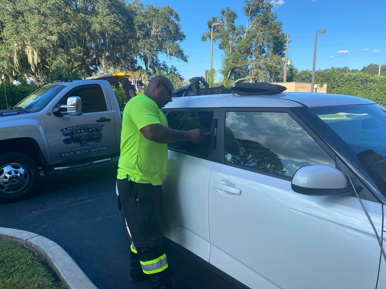 Worker in neon yellow shirt inspecting the window of a white car beside a truck outdoors.