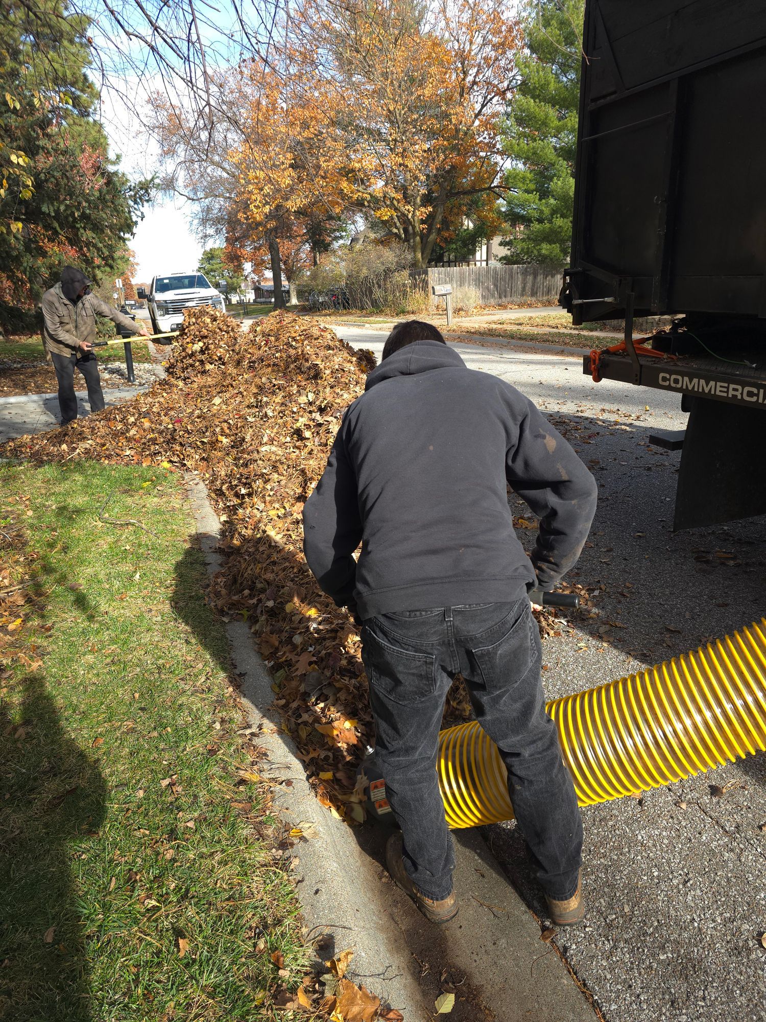 Workers clearing a leafy roadside drain beside a truck on a sunny autumn day