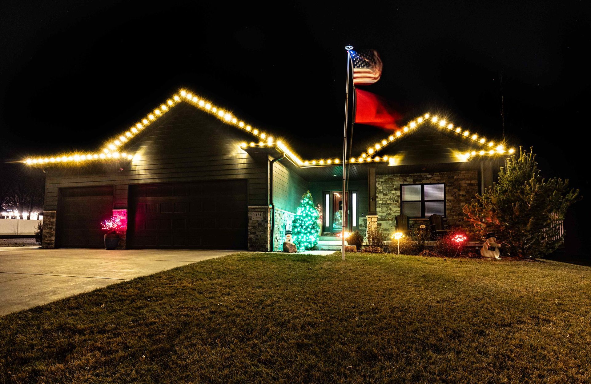 House lit up with Christmas lights at night, American flag flying, holiday decorations.