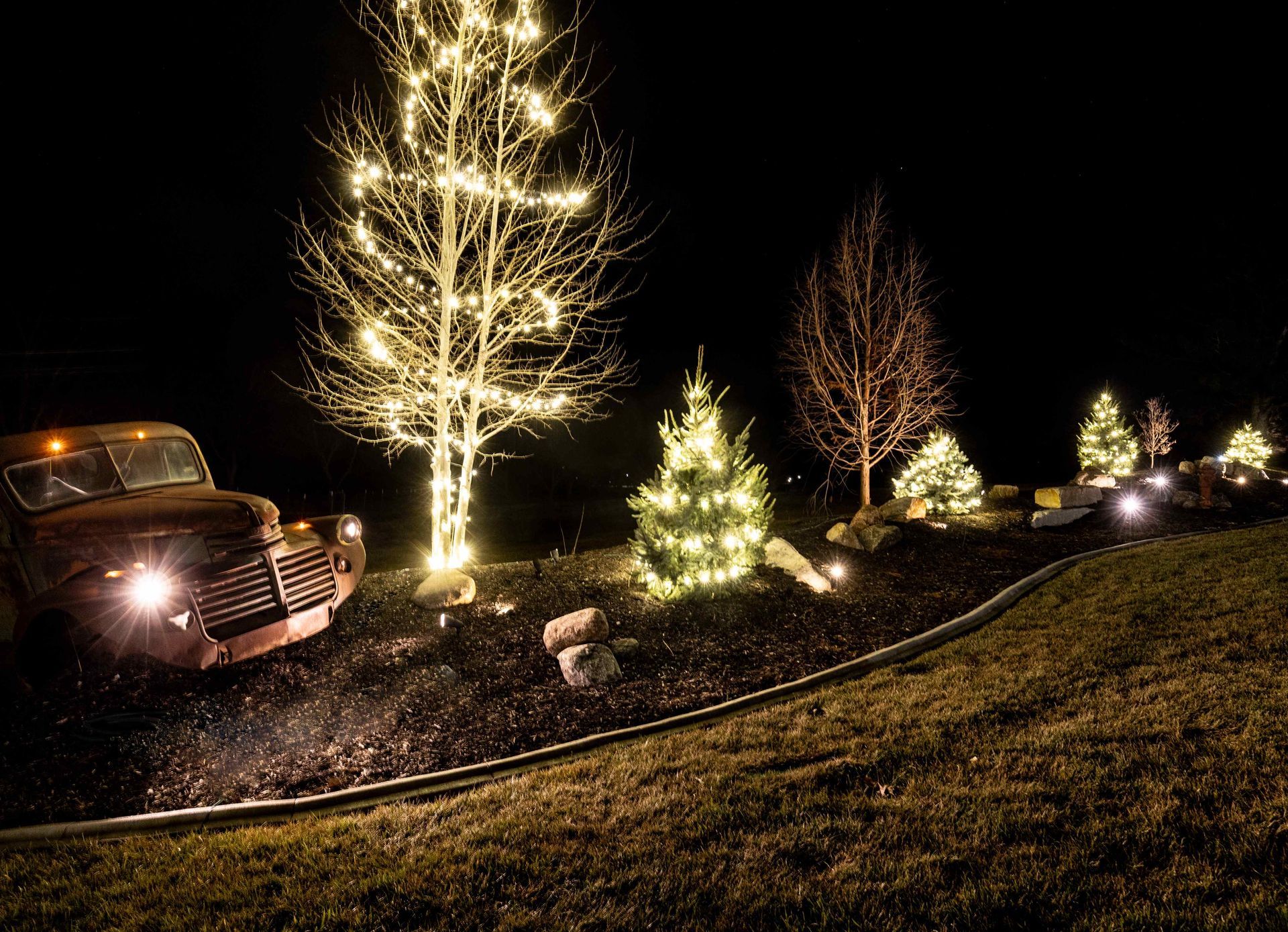 Old truck with headlights on, illuminated trees, Christmas lights, and dark night setting.