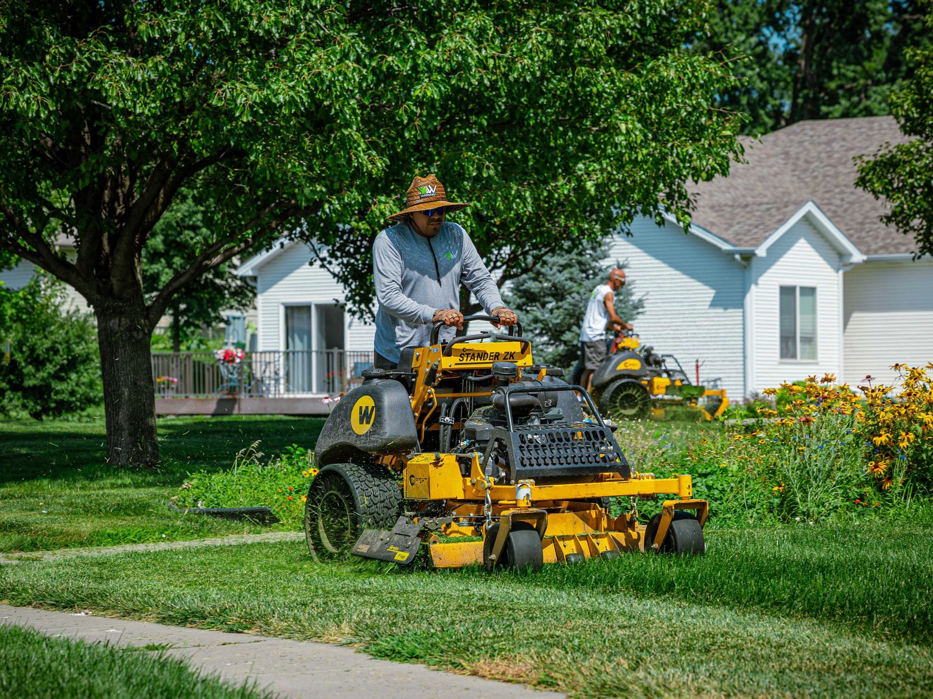 Worker mowing a suburban lawn with a yellow riding mower beside a white house